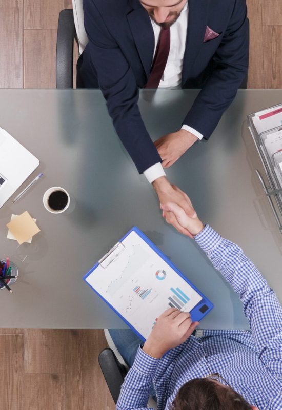 Top view of businessmen shaking hands during business negotiation in startup office discussing company strategy. Partners analyzing management profit planning investments meeting. Marketing documents